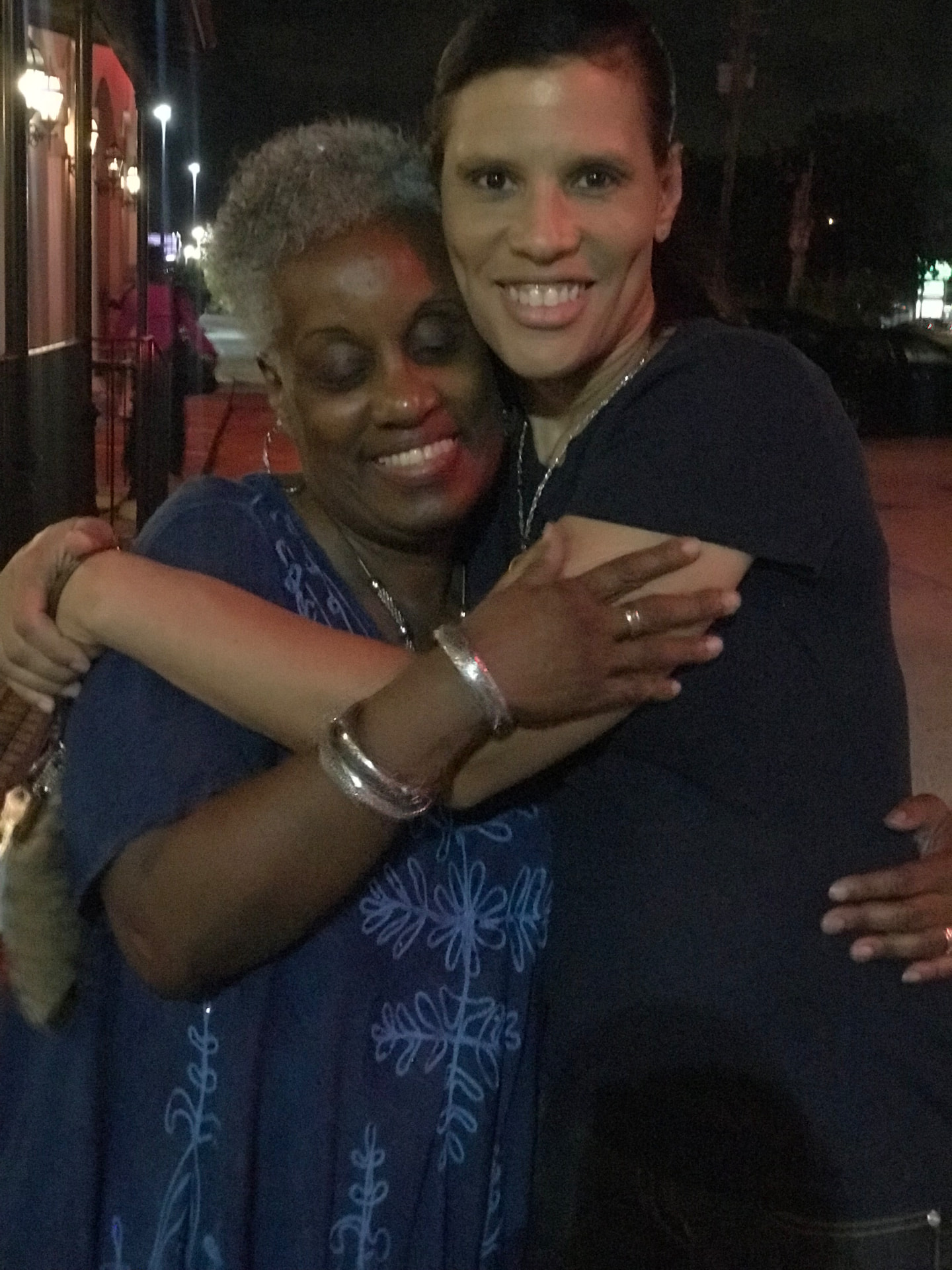 Momma Peggy holding a loved one close on a sidewalk at night, both smiling.