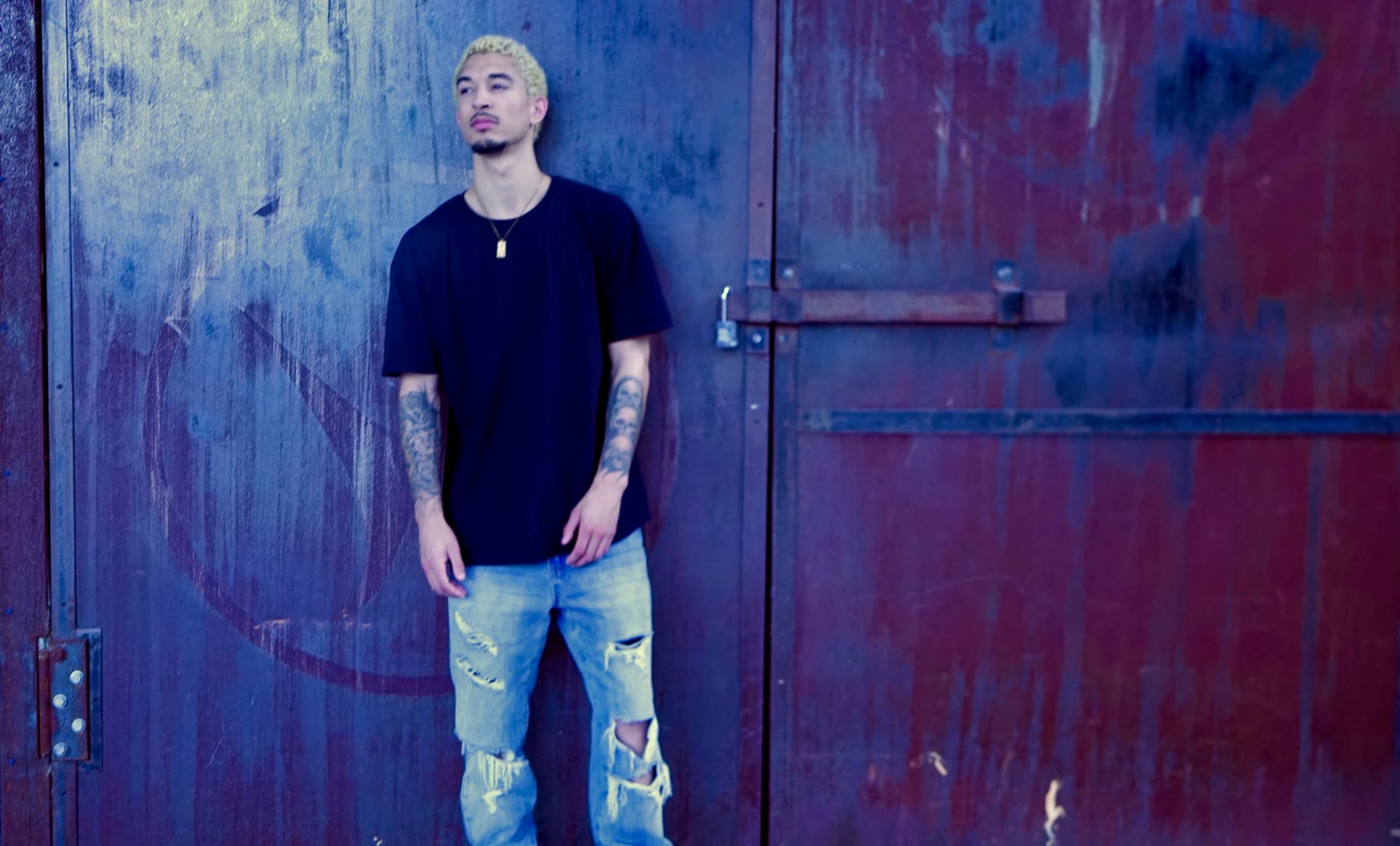 DUS10 photographed against a rusted industrial door in New Orleans, wearing an oversized black tee and distressed denim