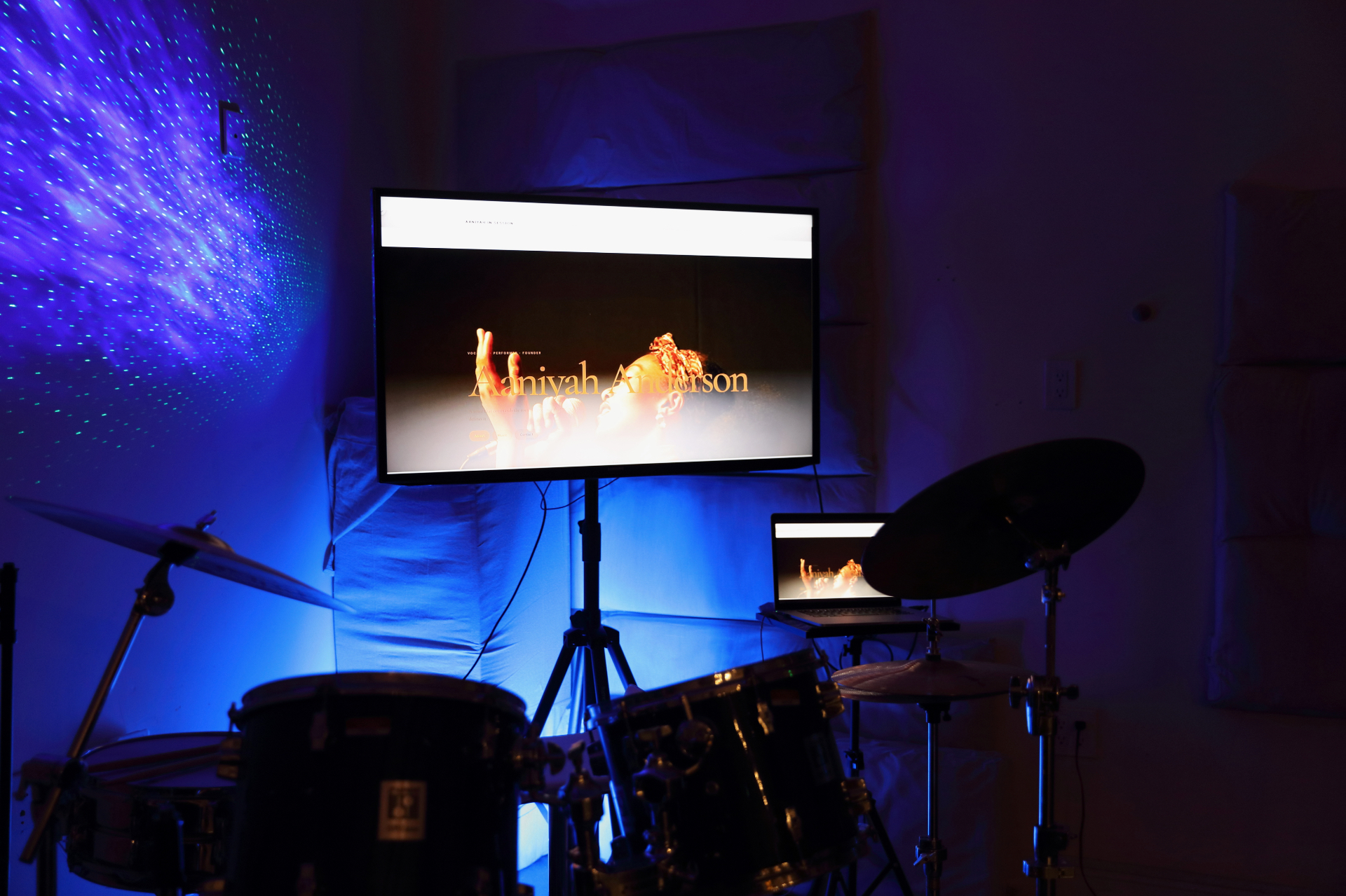 Aaniyah Anderson · site cut — Screening the Aaniyah Anderson site on the live-room TV, drum kit lit blue.