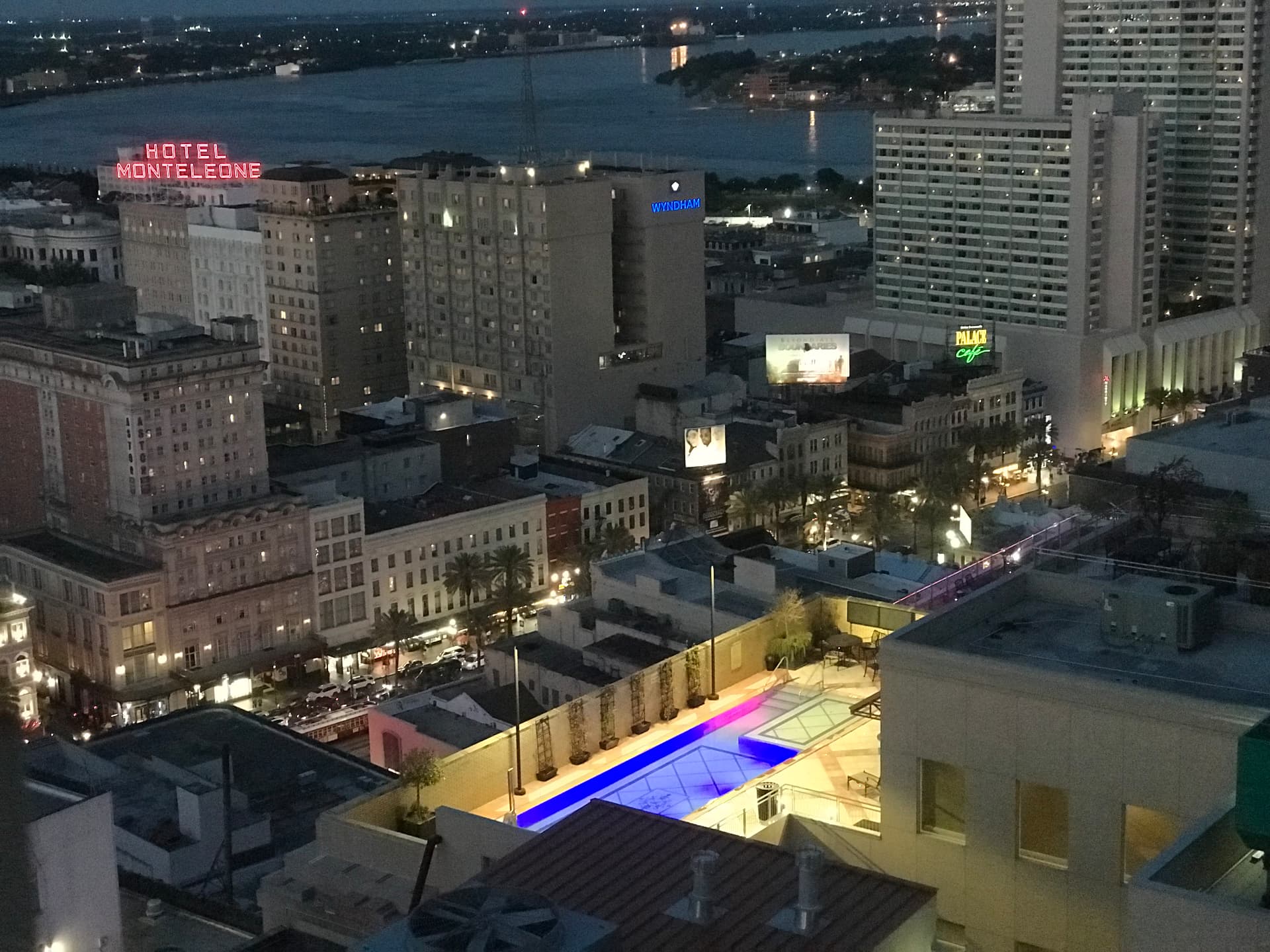 Blue-hour aerial view of the French Quarter with the Hotel Monteleone neon sign, Wyndham and Palace Café signage, and a violet-lit rooftop pool above the Mississippi River