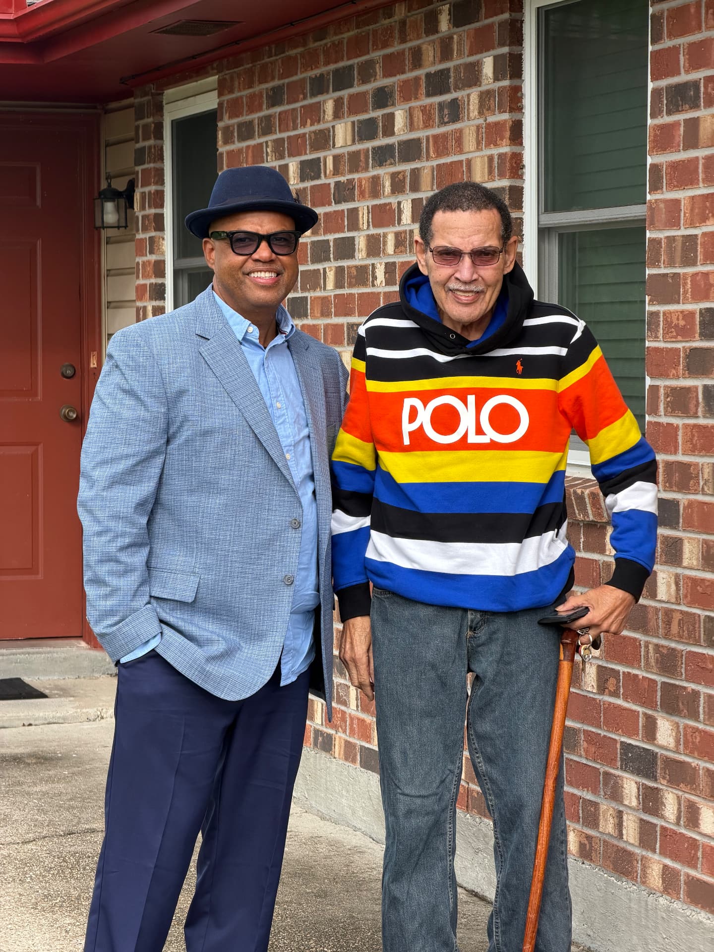 Byron Jupiter Jr. and his father, Byron Sr., standing together in front of a brick house. Byron Sr. is on the right.