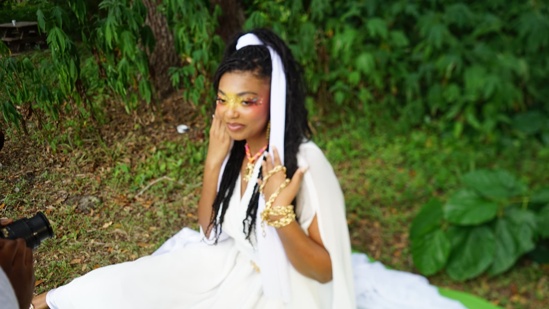 Aaniyah in white with gold face art and gold cuffs, hand raised to her cheek