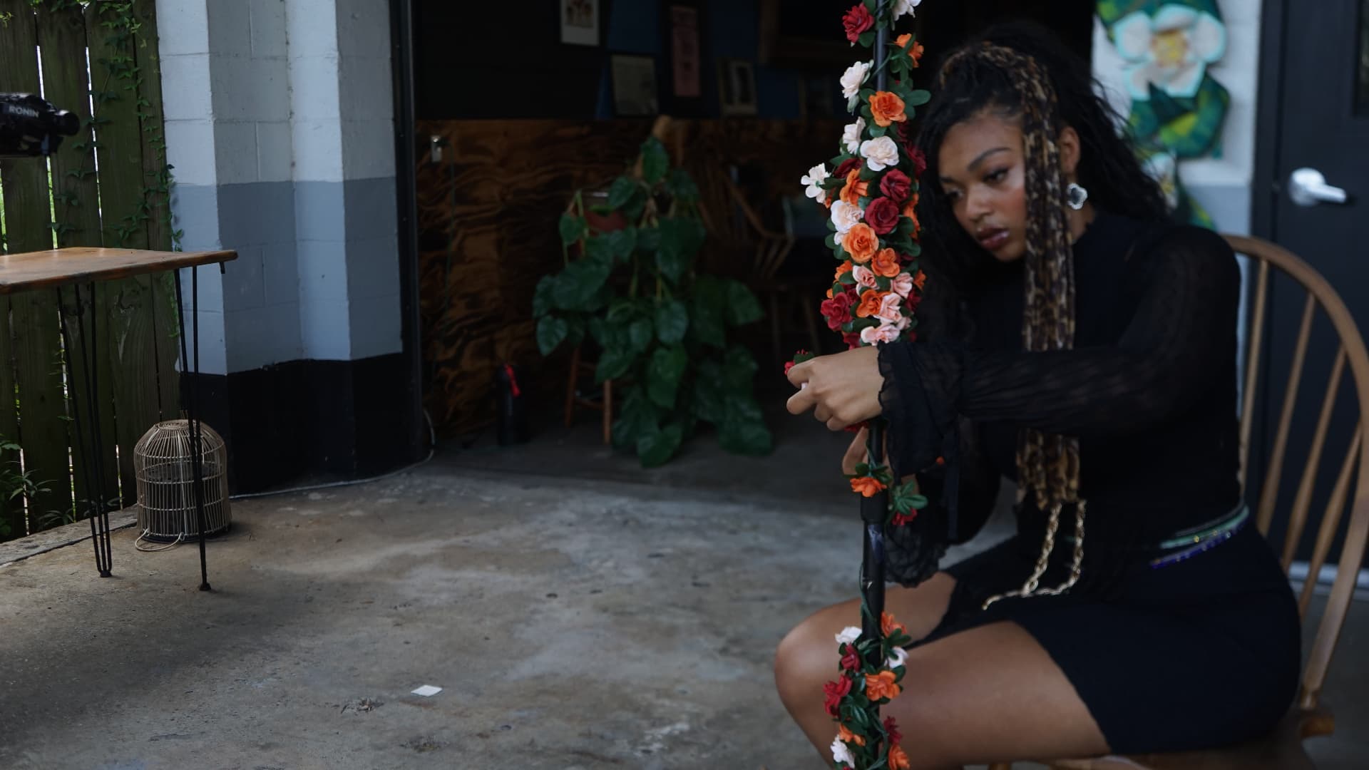 Aaniyah Anderson kneeling beside a flower-wrapped microphone stand on the patio during the Have It Your Way video shoot in New Orleans