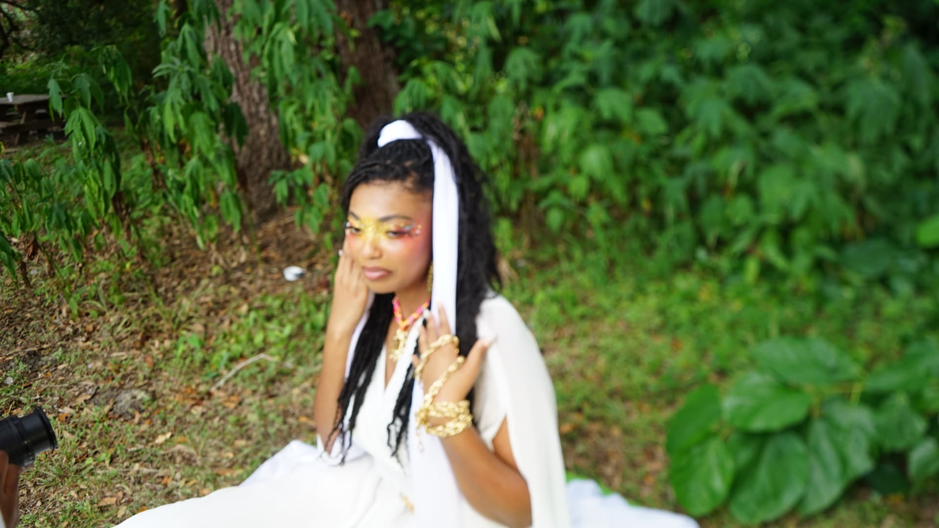 Aaniyah looking down, gold bracelets stacked on her wrist, greenery behind her