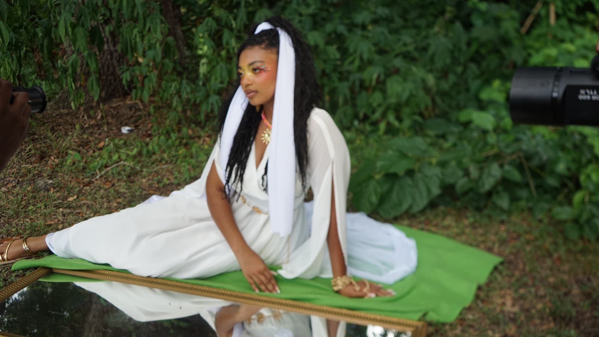Aaniyah seated in white on a green cloth beside a gilt mirror, cameras framing her, August 10, 2026