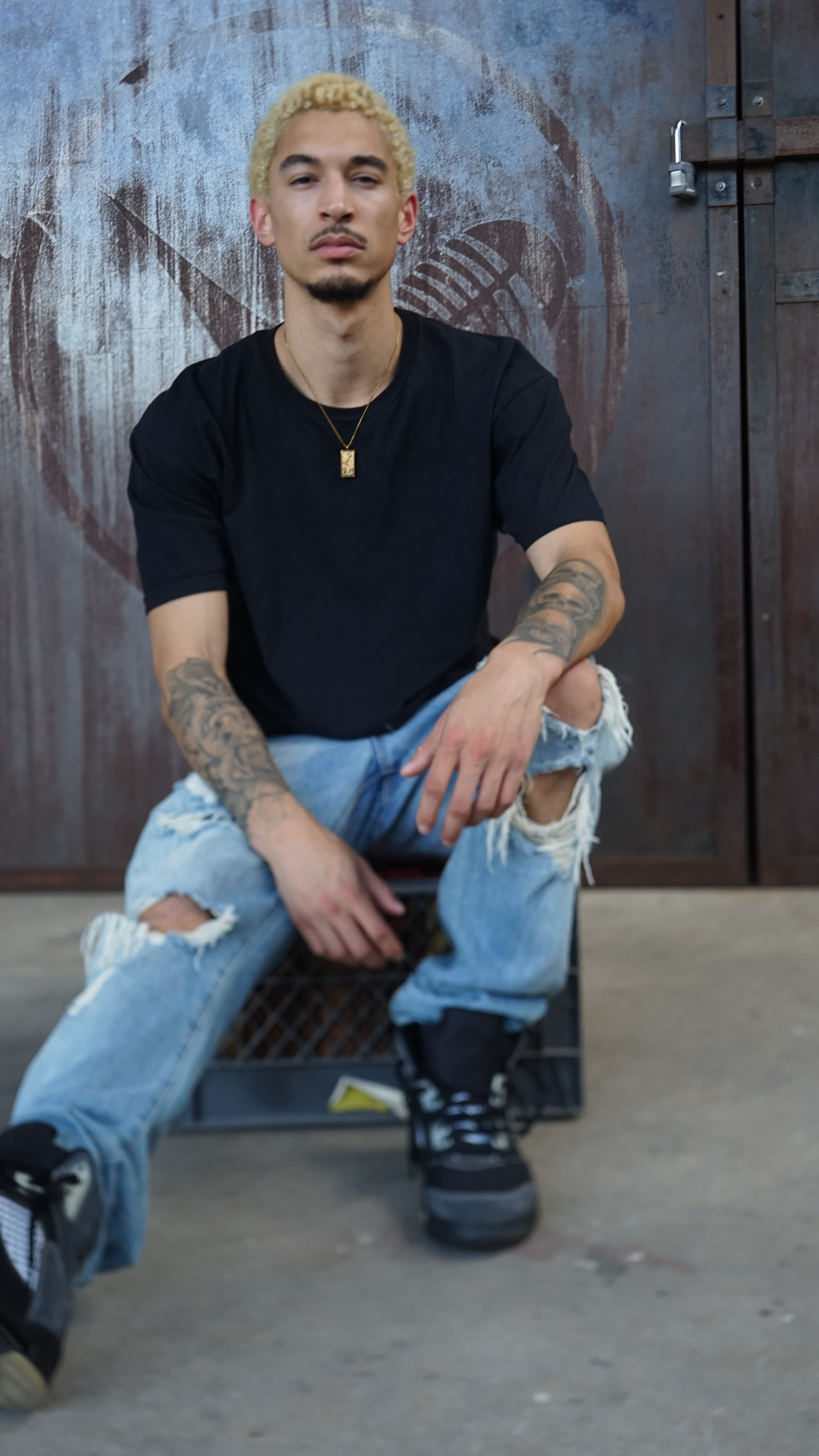 DUS10 seated on a wooden crate showing the denim stack over black high-top sneakers