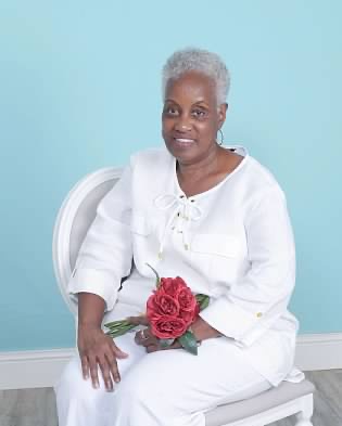 Portrait of Momma Peggy Jupiter seated in a white chair holding red roses, dressed in white against a soft blue backdrop.