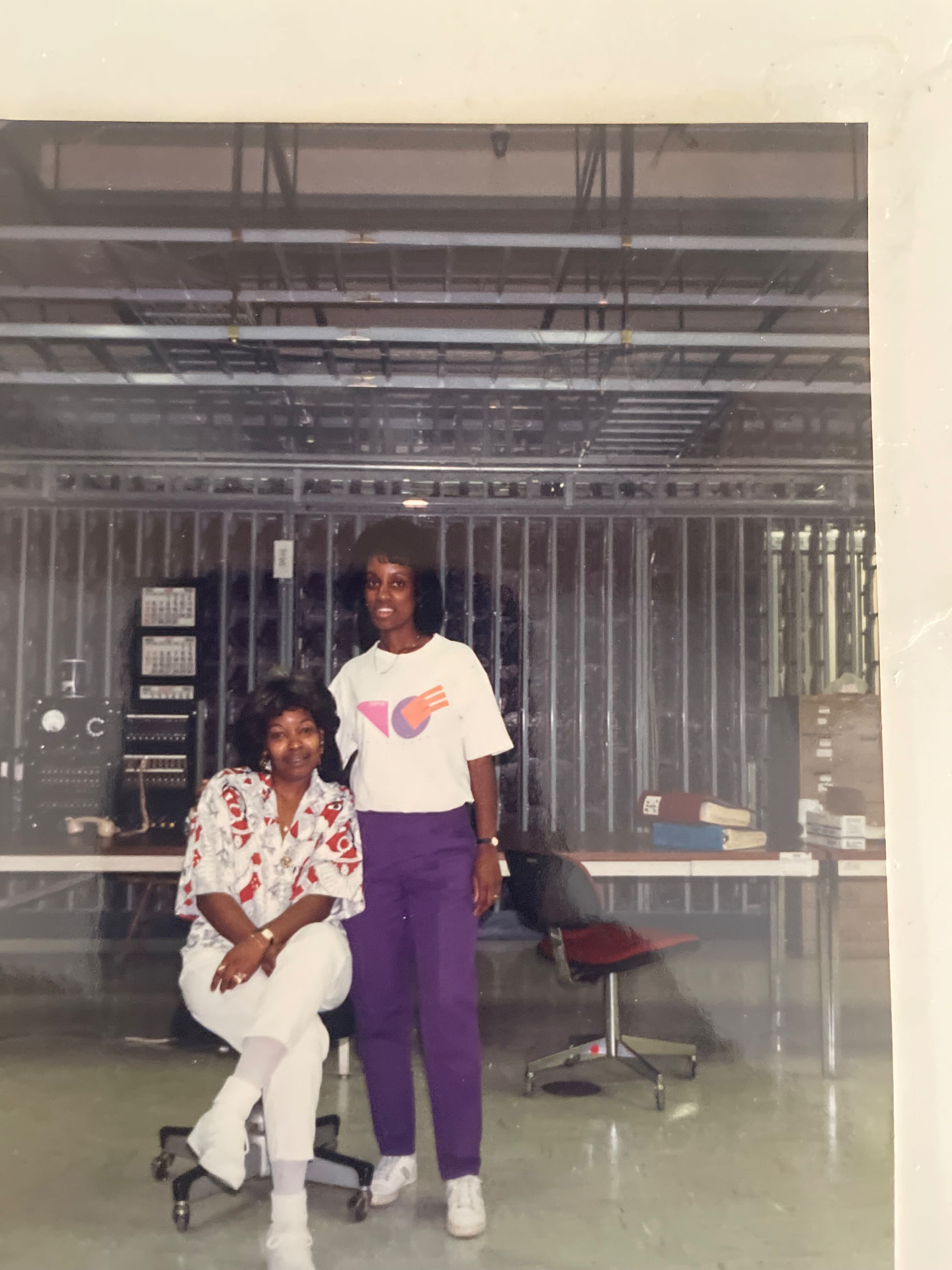 Momma Peggy seated at a work station in front of a telephone switchboard and equipment frames, with a coworker standing beside her.