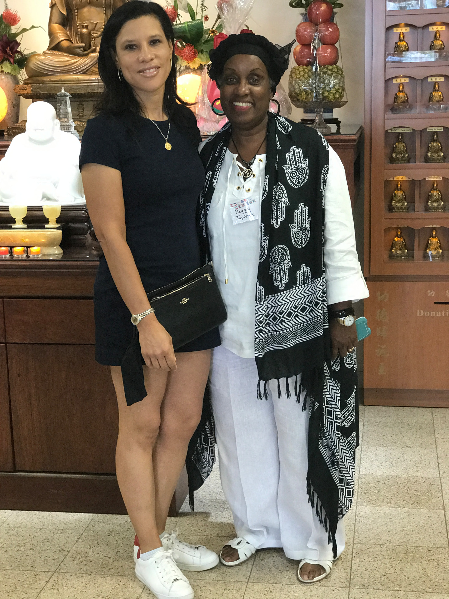 Momma Peggy in white linen and a black-and-white patterned scarf standing with a friend inside a temple, gilded statues and offerings behind them.